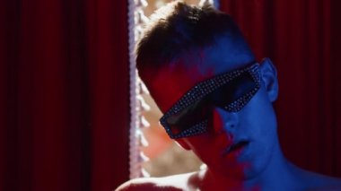 Güneş gözlükleriyle kaplı genç striptizci kırmızı perdelerin arkasında tuhaf danslar yapıyor.