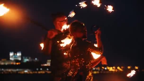 Fire show performance - usmívající se muž a žena v lesklém oblečení tančící s ohňovými pochodněmi na noční pláži — Stock video