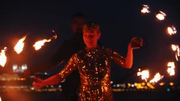Fire show performance - usmívající se žena a muž v lesklém oblečení tančící s ohňovými pochodněmi na noční pláži — Stock video