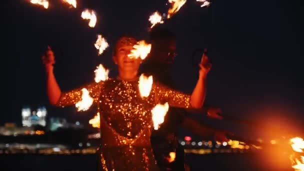 Ohňová show - žena a muž v lesklých šatech tančící s ohňovými pochodněmi na noční pláži — Stock video