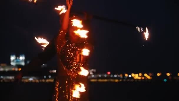 Fire show - žena v lesklých šatech a muž za ní tančí s ohňovými pochodněmi na noční pláži — Stock video