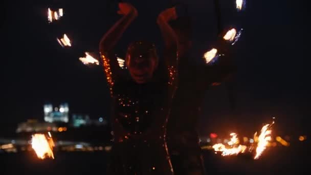 Фаер-шоу - улыбающаяся женщина в блестящем платье и мужчина за ее спиной танцуют с огненными факелами на ночном пляже — стоковое видео