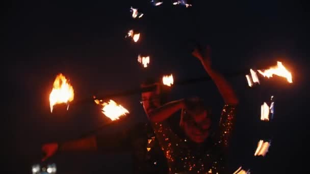 Fire show - usmívající se žena v lesklých šatech a muž za ní tančí s ohněm pochodně na noční pláži na pozadí města — Stock video