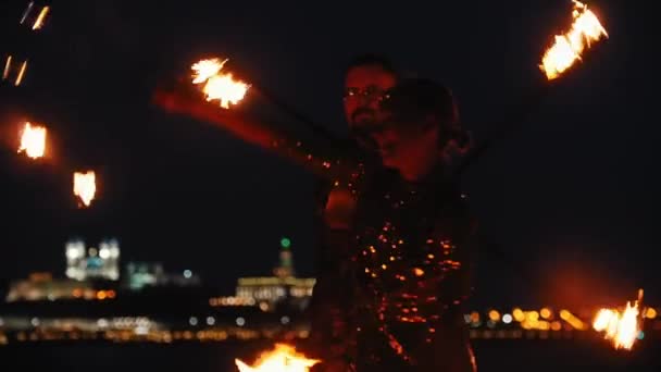 Fire show - usmívající se žena v lesklých šatech a muž za ní tančí s ohněm pochodně na pláži s noční město pozadí — Stock video