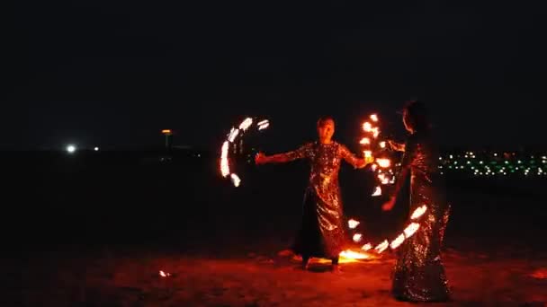 Fire show - two women in shiny dresses synchronously dancing with lit torches on the beach at night — Stock Video