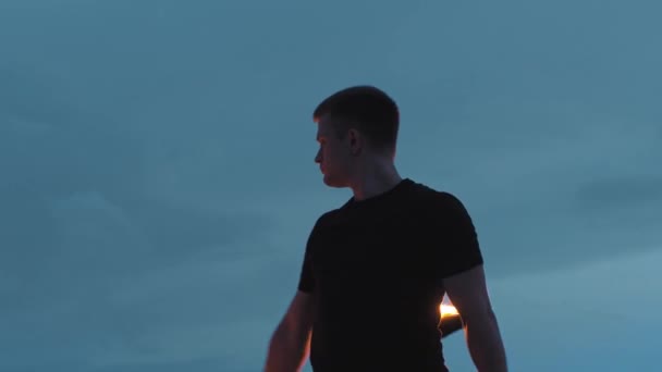 Jongeman performer in zwarte kleren draaien een vuur fakkel in zijn handen op een achtergrond van donkerblauwe lucht — Stockvideo