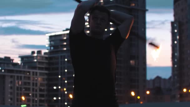 Jeune homme interprète en vêtements noirs tordant une torche de feu dans ses mains sur un fond de bâtiments modernes en fin de soirée — Video