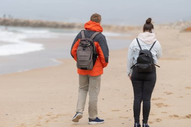 Sırt çantalı iki kişi okyanus kıyısındaki kumsalda yürüyorlar.