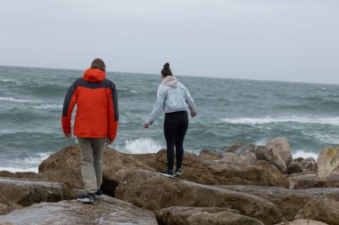 İki insan bulutlu bir gökyüzünün altında dalgalı denizde yürüyor.