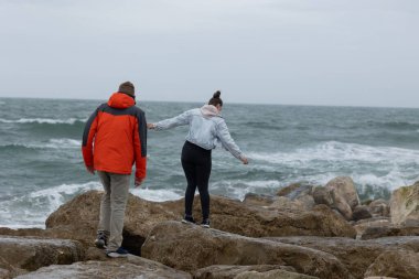 Genç çift dalgalı okyanusta kayaları keşfediyor, istikrar için el ele tutuşuyorlar.