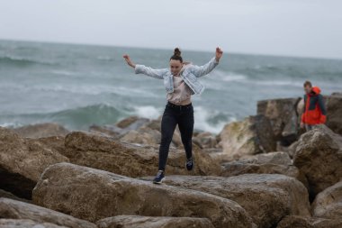 Genç bir kadın, arkadaşı izlerken okyanus dalgalarının yanındaki kayalarda dengede duruyor.