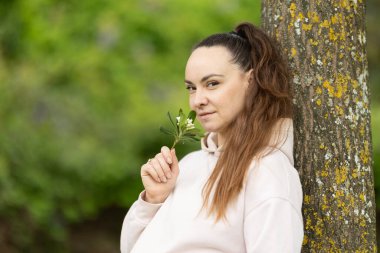 Elinde çiçek açan küçük bir dal tutan bir kadın, bir ağacın gövdesine yaslanıyor.