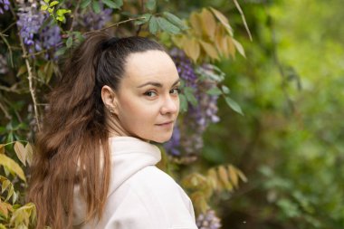 Kadın başını omzunun üstünden çeviriyor, çiçek açan Wisteria bahçesindeki kameraya bakıyor.