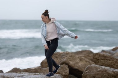 Sahildeki kayalıklarda dengede duran bir kadın ve arka planda okyanus dalgaları bulutlu bir gökyüzünün altında.