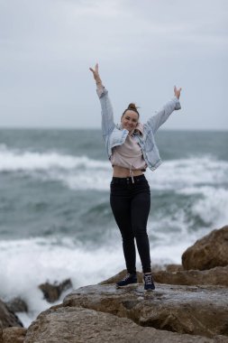 Kolları havada kayaların üzerinde duran genç bir kadın, deniz kenarında mutlulukla gülümsüyor.