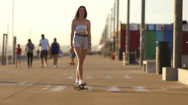 Gün batımında deniz kenarında bir sahil yolunda kameraya doğru kayan kendinden emin genç bir kadın.