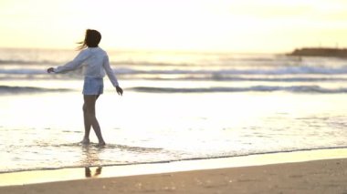 Mutlu genç kadın deniz kıyısında geziniyor, altın saat ışığının ve nazik dalgaların tadını çıkarıyor
