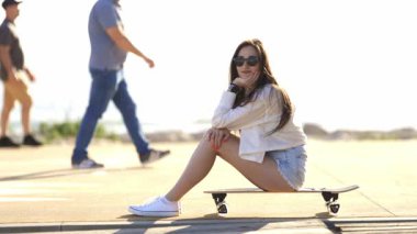 İnsanlar arka planda yürürken, güneş gözlüklü çekici genç bir kadın kaykayında oturuyor.