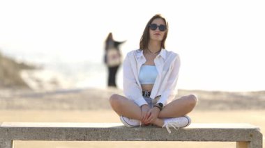 Güzel genç bir kadın kumsalda bankta oturmuş güneşli bir günün tadını çıkarıyor.