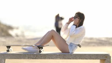 Güzel genç bir kadın beton bir bankta kaykayıyla güneşli bir günün tadını çıkarıyor.