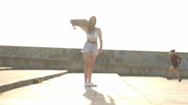 Kaykaylı neşeli genç bir kadın güneşli bir şehirde köpeğiyle mutlu bir şekilde dans ediyor.