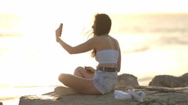 Gün batımında deniz kenarındaki bir kayanın üzerinde oturan genç bir kadın akıllı telefonuyla selfie çekiyor.