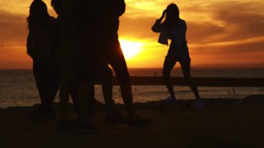 Yürüyen insanların siluetleri ve deniz kıyısındaki altın saatin tadını çıkaran kaykaylı genç bir kadın.