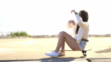 Güzel genç bir kadın kaykayın üzerinde oturuyor ve güneşli bir deniz kenarında geziniyor.