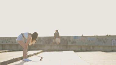 Çekici genç bir kadın uzun bir tahtaya biniyor ve gün batımında beton bir set boyunca sürüyor.