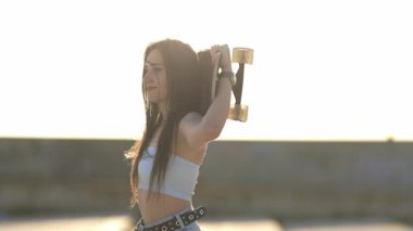 Güzel bayan kaykaycı kafasının arkasında uzun bir sörf tahtası tutarken altın saatin tadını çıkarıyor.