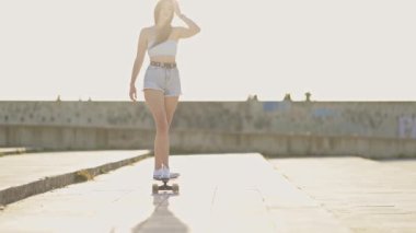 Güneşli bir günde beton bir gezinti güvertesinde kaykay süren çekici genç bir kadın.