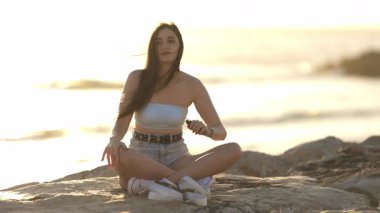 Genç bir kadın kayalık bir sahilde bacak bacak üstüne atmış ve gün batımında elektronik bir sigarayla buharlaşıyor.