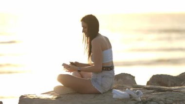 Arka planda okyanus manzaralı cep telefonunu kullanarak kıyı kayalarında oturan arka ışıkçı kadın.