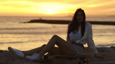 Kumsalda uzun tahtasında oturan güzel genç bir kadın rahatlatıcı altın bir günbatımının tadını çıkarıyor.