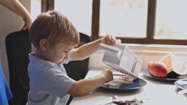 Sevimli çocuk güneşli bir sabahta mutfak masasında yemek pişirmeyi ve rendelemeyi öğreniyor.