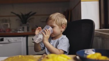 Sevimli küçük sarışın çocuk su içiyor ve mutfak masasında otururken yaramazlık yapıyor.