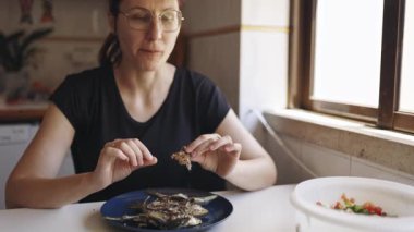 Mutfak masasında oturan bir kadın kızarmış balık yiyor, kemikleri filetodan ayırıyor.