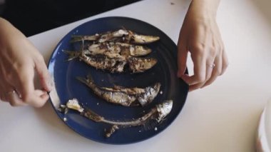 Kadınların elleri titizlikle beyaz bir masada koyu mavi bir tabakta küçük ızgara balığın kemiklerini çıkarıyor.