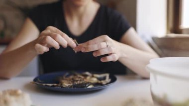 Gözlüklü kadın mutfaktaki masada oturuyor, elleriyle ızgara balık yiyor.