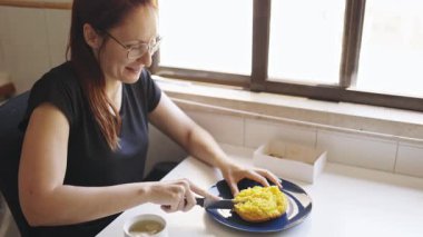 Gözlüklü, gülümseyen kadın kahvaltıda pencerenin yanında sarı mısır keki kesiyor.