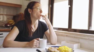 Genç bir kadın sabahları evde kahve içerken bir tabak omlet ve et yiyor.