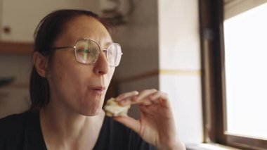 Orta yaşlı, gözlüklü bir kadın mutfaktaki pencerenin yanında büyük bir zevkle sandviç yiyor.