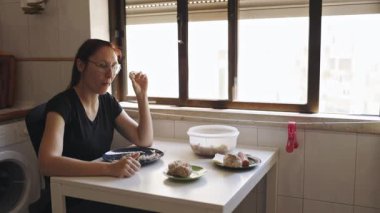 Gözlüklü yalnız bir kadın pencerenin yanındaki küçük bir mutfak masasında tek başına yemek yiyor.