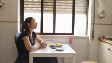 Gözlüklü, düşünceli bir kadın. Masada tek başına oturmuş, pencerenin kenarında kahvaltı yapıyor.