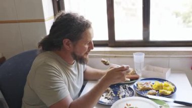 Uzun saçlı, sakallı bir adam bir masada oturmuş mutfağındaki pencerenin kenarında yemek yiyor.