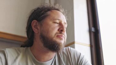 Sakallı, uzun saçlı, yemek yiyen ve pencereden dışarı bakan dalgın bir adam.