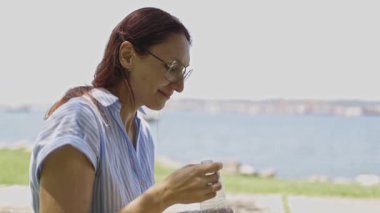 Gözlüklü bir kadın güneşli bir günde dışarıda oturuyor plastik bir şişeden tatlı su içiyor.