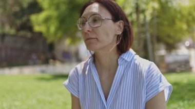 Şehir parkında yeşil çimlerde oturan gözlüklü orta yaşlı bir kadın sağlıklı bir şeyler yiyor.