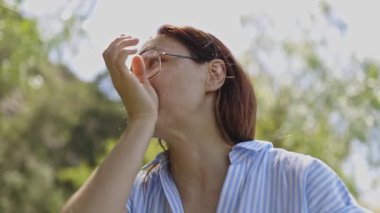 Parkta güneşli bir günde sulu bir elma yiyen gözlüklü kızıl saçlı kadın.