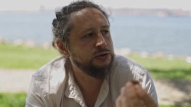 Orta yaşlı sakallı bir adam ve güneşli bir günde deniz kenarında bir parkta atıştırmalık yiyen bir adam.